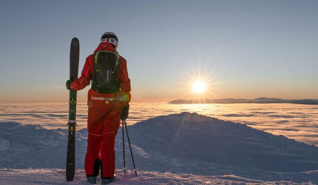 Man in a red outfit holding skis and poles, enjoying a beautiful sunset in the mountains